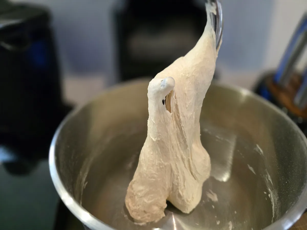 Pizzateig hängt am Knethaken der Küchenmaschine — sichtbar ausgeprägtes Glutennetz durch Dehnung